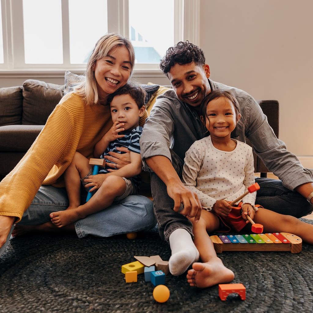 Two adults and two children sitting on the floor in a cozy living room, playing with colorful building blocks and a wooden toy xylophone.